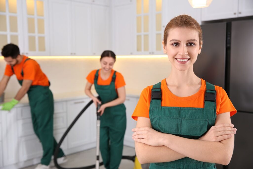 3 professional cleaners in green overalls deep-cleaning a kitchen while a young woman smiles.