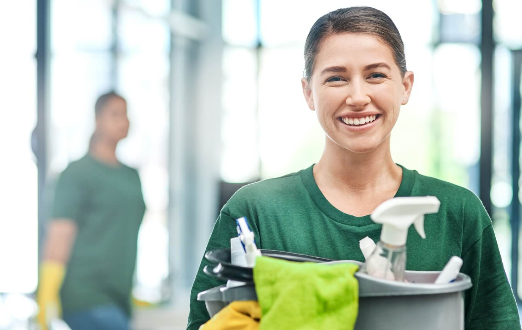 A professional cleaner wearing a green shirt holds a bucket full of cleaning supplies and clothes and smiles brightly.