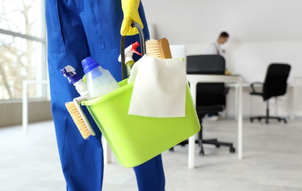 A professional cleaner carries a green bucket. The bucket holds a variety of brushes, cleaning supplies, and towels.