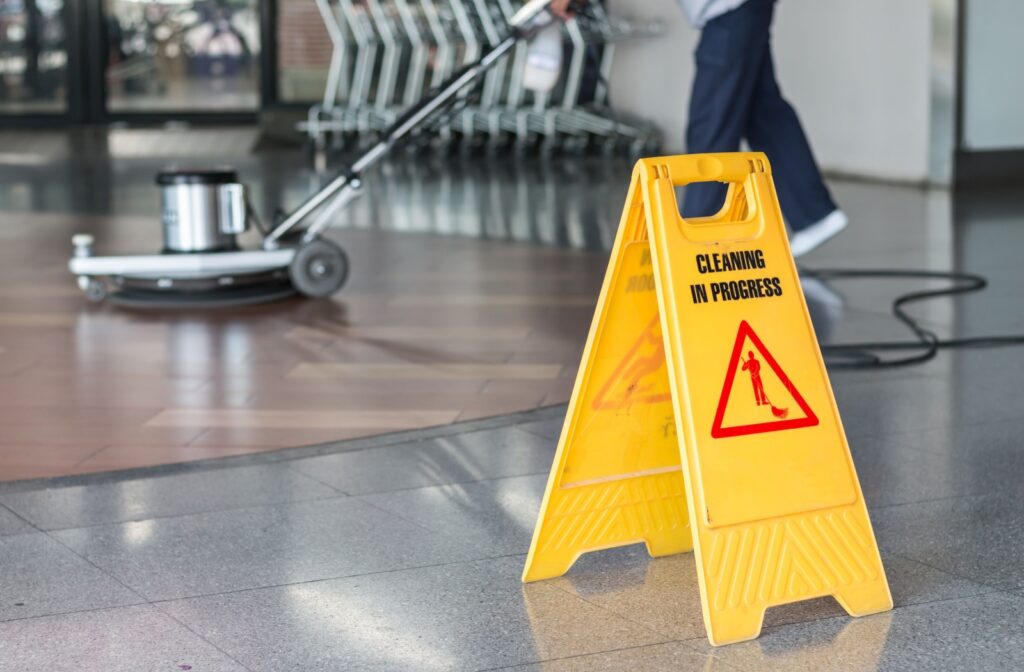 An image of a cleaner polishing an office floor with a floor polishing machine next to a sign that reads "cleaning in progress".