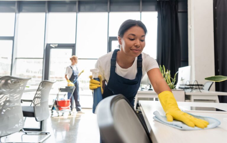 A professional cleaner wearing yellow rubber gloves finishes cleaning a desk in a bright, clean office space.