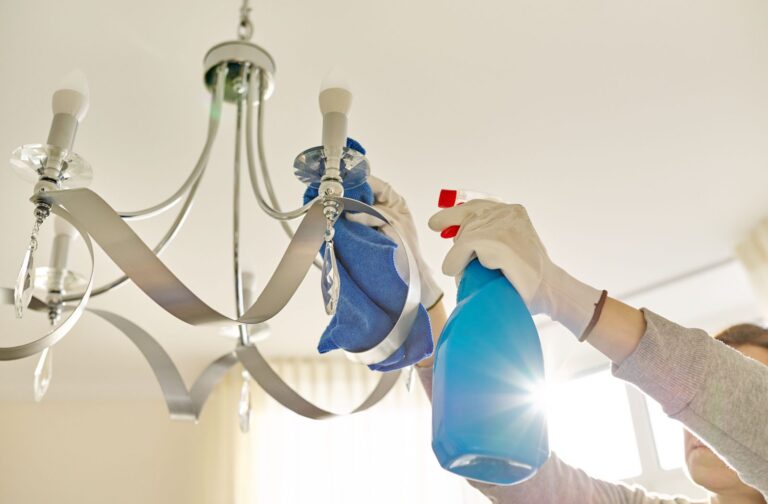A person's hands in white gloves reaching up to clean a chandelier with a blue spray bottle and blue cloth.
