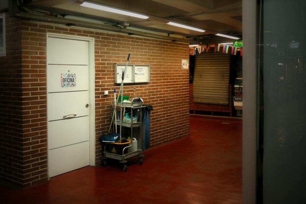 A brightly lit industrial hallway with a cleaning cart and supplies near an office door.